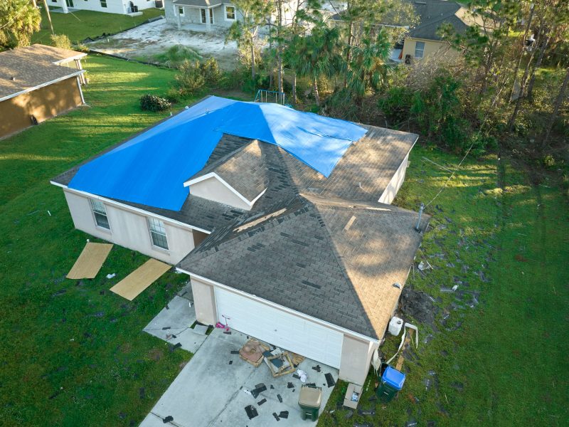 Aerial view of damaged in hurricane Ian house roof covered with blue protective tarp against rain water leaking until replacement of asphalt shingles.